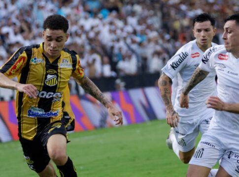 Um jogador de futebol veste o uniforme listrado em amarelo e preto do Grêmio Novorizontino enquanto conduz a bola em velocidade pelo gramado. Ele é perseguido de perto por dois jogadores do Santos FC, que vestem uniformes brancos. Ao fundo, a arquibancada está repleta de torcedores, e as placas de publicidade coloridas margeiam o campo. A ação ocorre durante o dia sob iluminação natural.