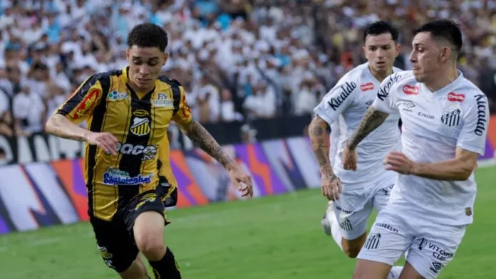 Um jogador de futebol veste o uniforme listrado em amarelo e preto do Grêmio Novorizontino enquanto conduz a bola em velocidade pelo gramado. Ele é perseguido de perto por dois jogadores do Santos FC, que vestem uniformes brancos. Ao fundo, a arquibancada está repleta de torcedores, e as placas de publicidade coloridas margeiam o campo. A ação ocorre durante o dia sob iluminação natural.