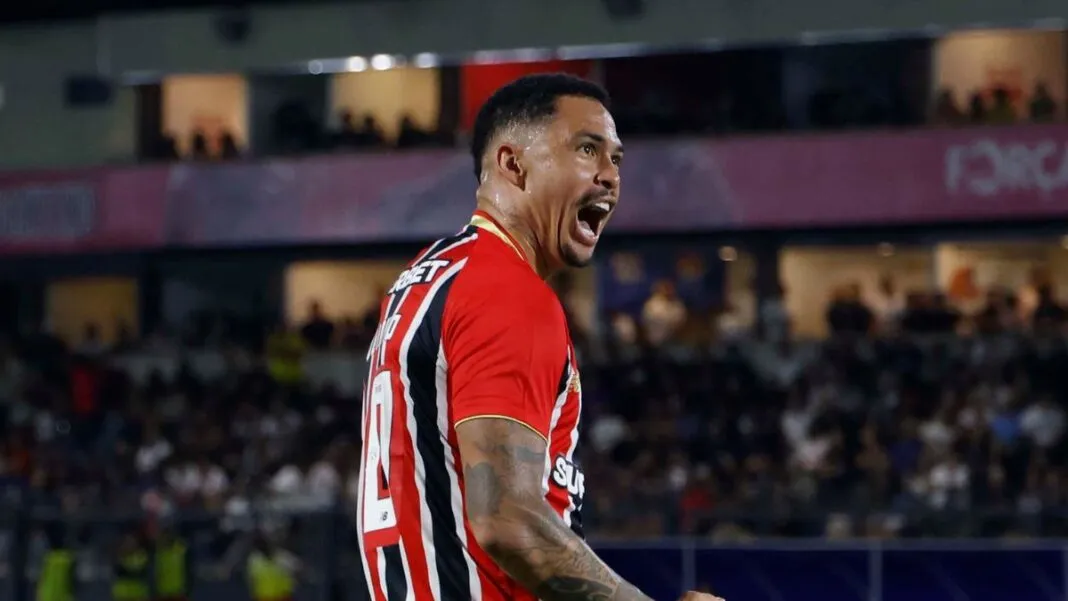 O jogador Luciano, do São Paulo FC, celebra intensamente em campo durante uma partida noturna. Ele está de perfil, com a boca aberta em um grito de comemoração e os punhos cerrados. Veste a camisa listrada em vermelho, branco e preto, calções brancos e meiões brancos. Ao fundo, as arquibancadas do estádio estão lotadas e iluminadas pelos refletores.