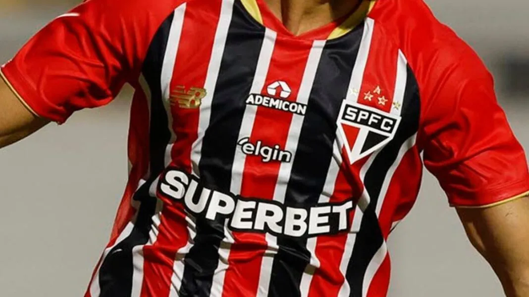 Close-up do tronco de um jogador vestindo a camisa listrada do São Paulo Futebol Clube, em vermelho, preto e branco. Diversos logotipos de patrocinadores estão visíveis no uniforme, incluindo o escudo do time no peito e marcas comerciais no centro e ombros.