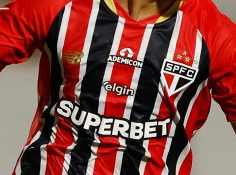 Close-up do tronco de um jogador vestindo a camisa listrada do São Paulo Futebol Clube, em vermelho, preto e branco. Diversos logotipos de patrocinadores estão visíveis no uniforme, incluindo o escudo do time no peito e marcas comerciais no centro e ombros.