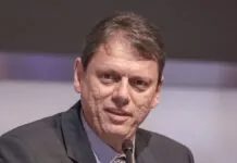 Um homem de pele clara e cabelos curtos castanhos aparece em um plano fechado, falando em um microfone que está posicionado logo abaixo de seu rosto. Ele veste um paletó escuro sobre uma camisa social azul clara e gravata estampada. Sua expressão é séria e focada. O fundo está desfocado em tons de cinza e lilás.