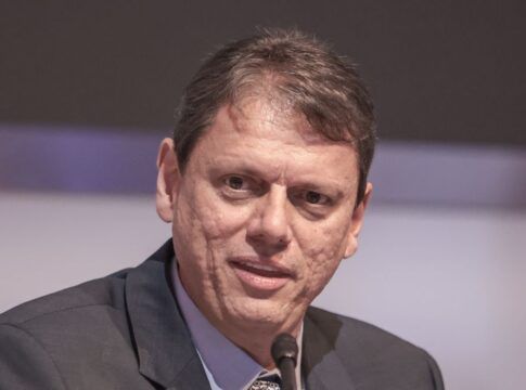 Um homem de pele clara e cabelos curtos castanhos aparece em um plano fechado, falando em um microfone que está posicionado logo abaixo de seu rosto. Ele veste um paletó escuro sobre uma camisa social azul clara e gravata estampada. Sua expressão é séria e focada. O fundo está desfocado em tons de cinza e lilás.