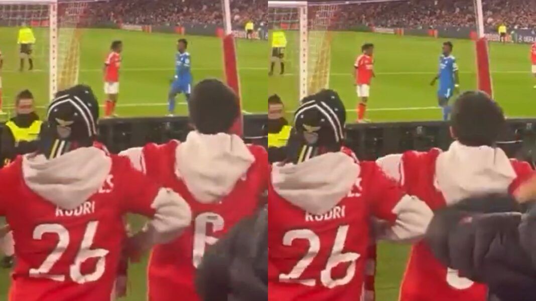 Montagem com duas imagens de baixa qualidade registradas da arquibancada de um estádio. Em primeiro plano, torcedores vestindo casacos vermelhos gesticulam em direção ao campo. No gramado, ao fundo, dois jogadores — um de uniforme vermelho e outro de azul — estão próximos.