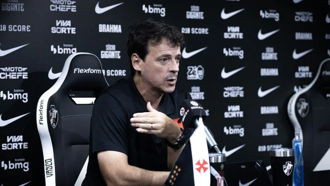 O técnico Fernando Diniz concede uma entrevista coletiva. Ele está sentado em uma cadeira esportiva preta e branca com o escudo do Vasco da Gama, vestindo uma camisa polo preta. Com a mão direita levemente levantada em um gesto explicativo, ele olha para a frente. À sua frente, sobre a mesa, há um microfone com a marca 