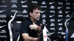O técnico Fernando Diniz concede uma entrevista coletiva. Ele está sentado em uma cadeira esportiva preta e branca com o escudo do Vasco da Gama, vestindo uma camisa polo preta. Com a mão direita levemente levantada em um gesto explicativo, ele olha para a frente. À sua frente, sobre a mesa, há um microfone com a marca 