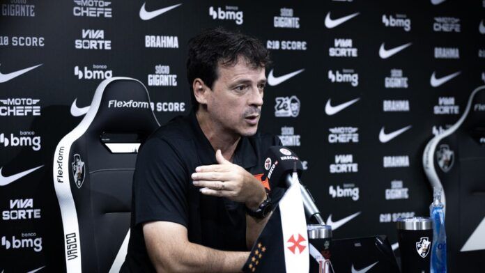 O técnico Fernando Diniz concede uma entrevista coletiva. Ele está sentado em uma cadeira esportiva preta e branca com o escudo do Vasco da Gama, vestindo uma camisa polo preta. Com a mão direita levemente levantada em um gesto explicativo, ele olha para a frente. À sua frente, sobre a mesa, há um microfone com a marca 