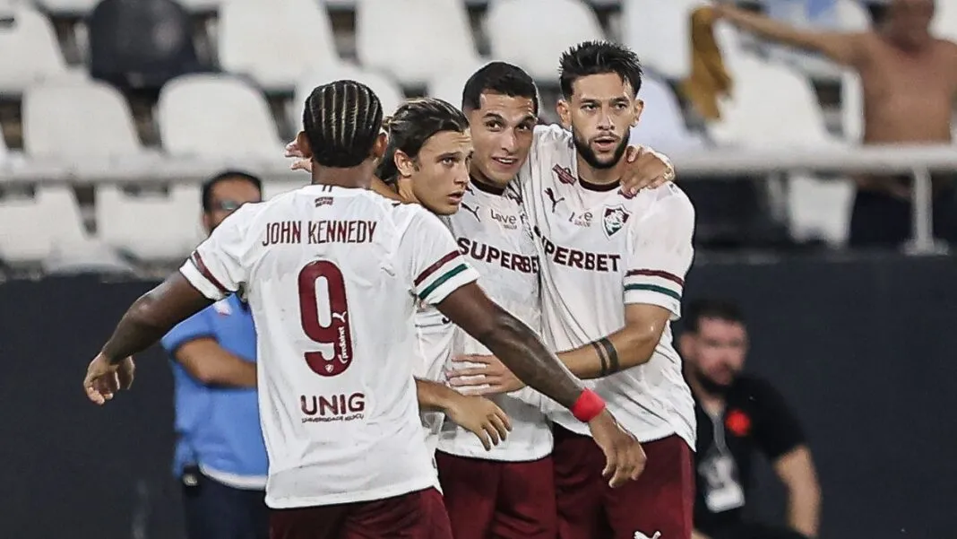 Quatro jogadores de futebol do Fluminense FC celebram juntos no gramado. Três deles estão de frente, abraçados, vestindo a camisa branca com detalhes em grená e verde e calções grenás. O quarto jogador está de costas, exibindo o nome 