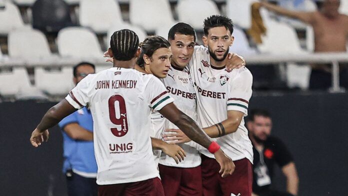 Quatro jogadores de futebol do Fluminense FC celebram juntos no gramado. Três deles estão de frente, abraçados, vestindo a camisa branca com detalhes em grená e verde e calções grenás. O quarto jogador está de costas, exibindo o nome 